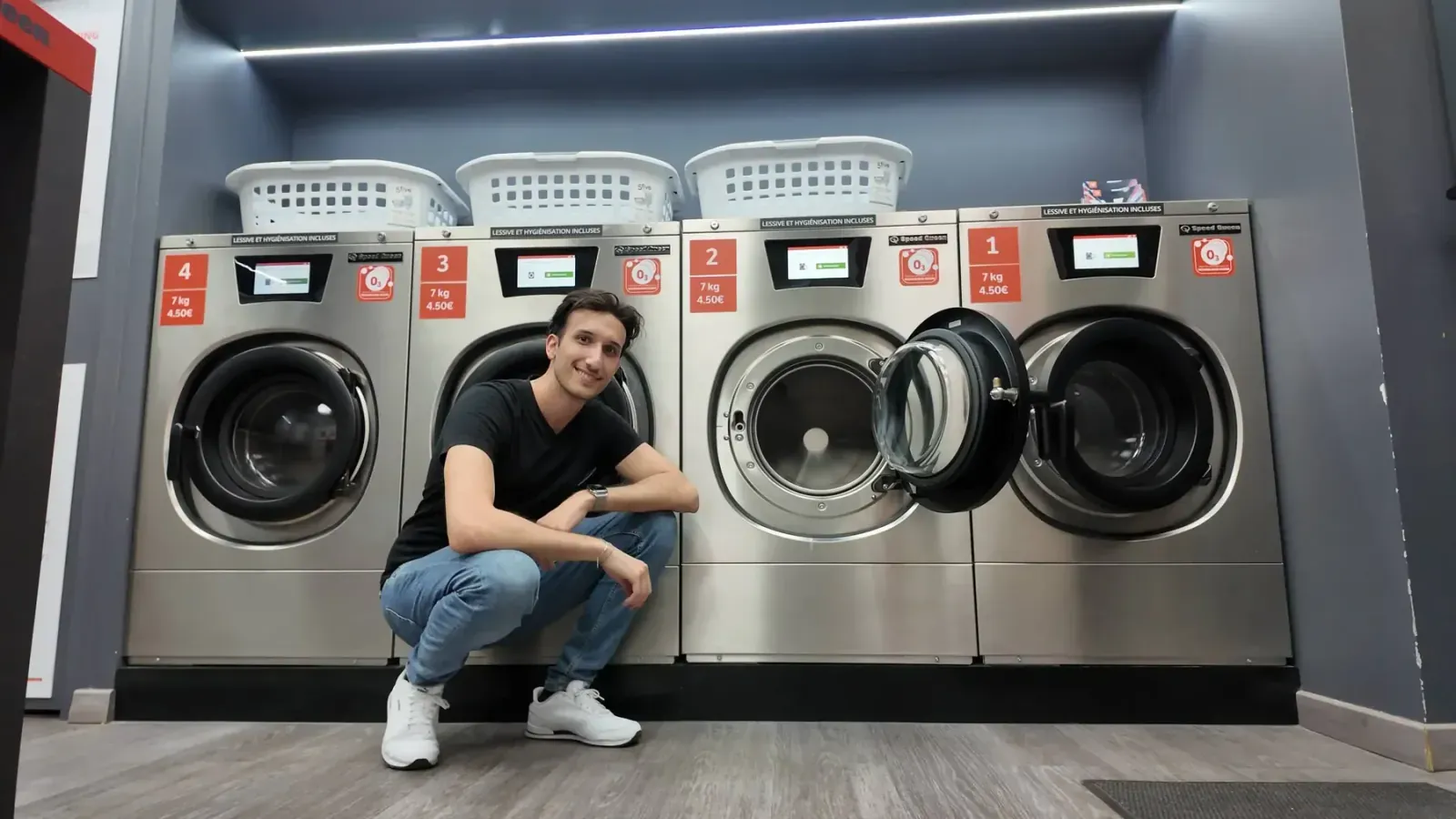 Maxime accroupi devant les quatre machines à laver 7 kg de la laverie, 3 Square Paul Paray, Paris 17ᵉ